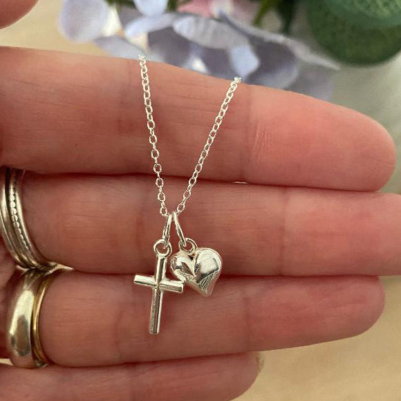 Silver necklace with cross and heart charm held in hand.