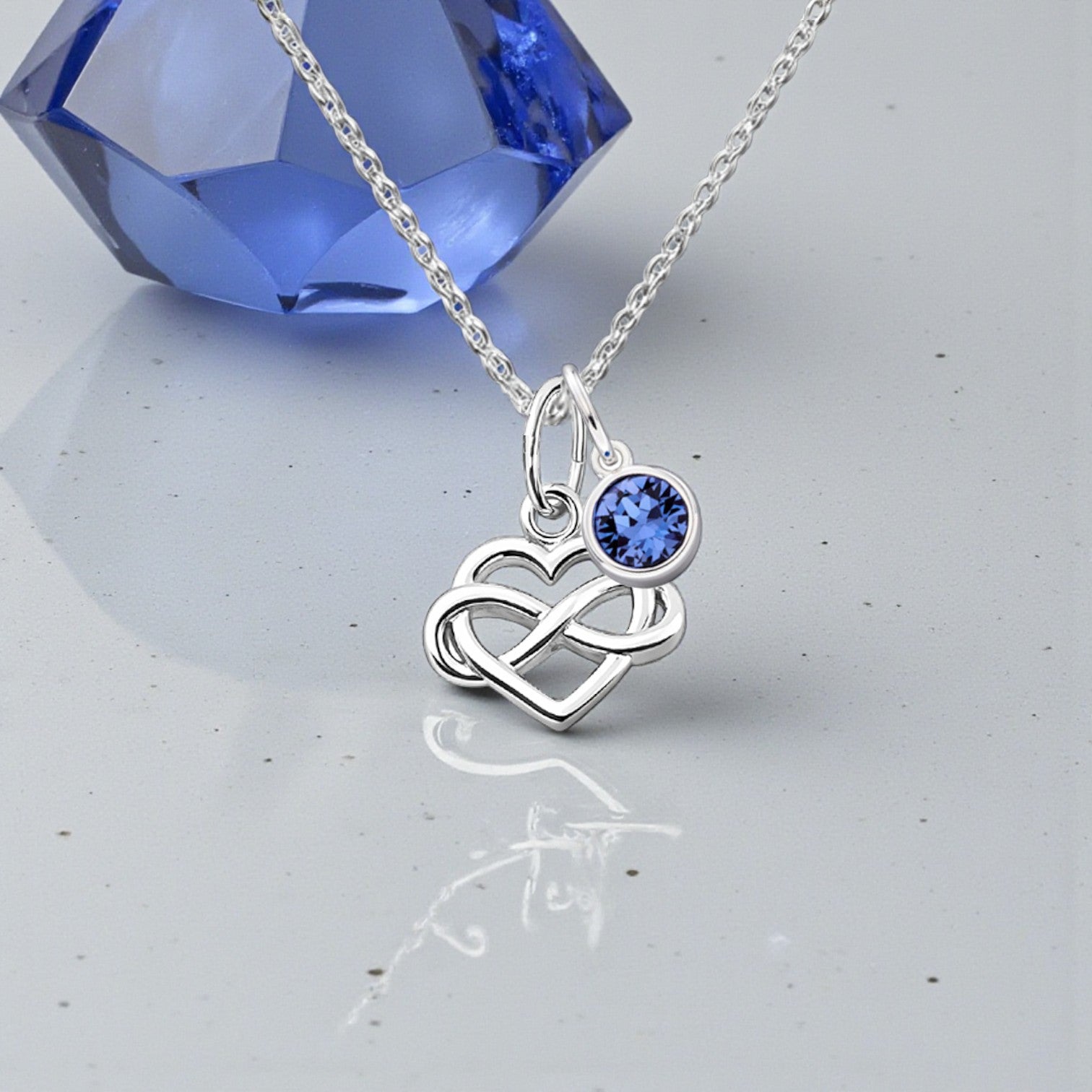 Silver necklace with a heart and infinity symbol pendant featuring a blue gemstone, set against a light gray background.