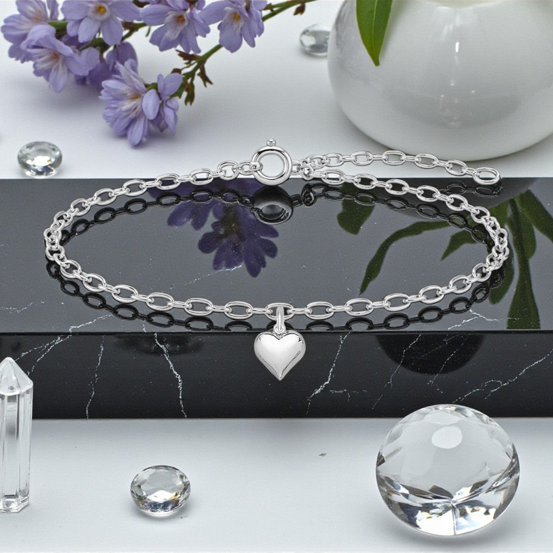 Silver bracelet with heart charm on a reflective surface with flowers and crystals in the background